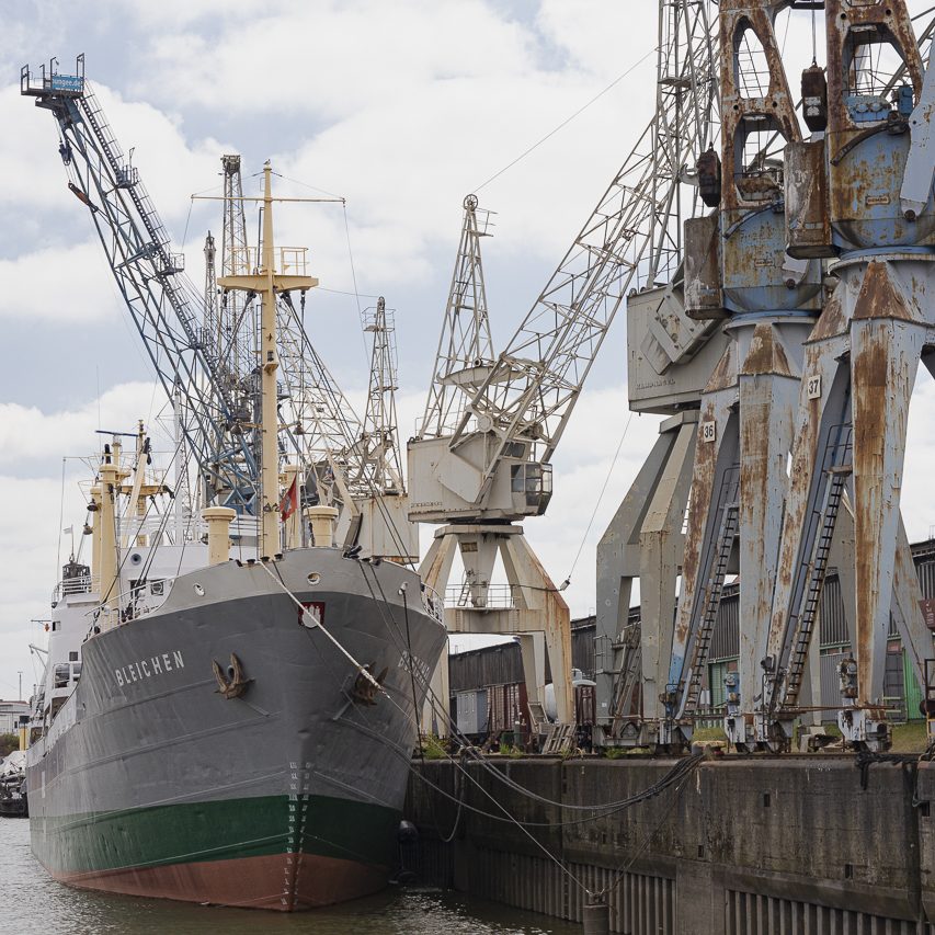 Ein Schiff im Hafen, umgeben von großen Kränen und industriellen Anlagen.