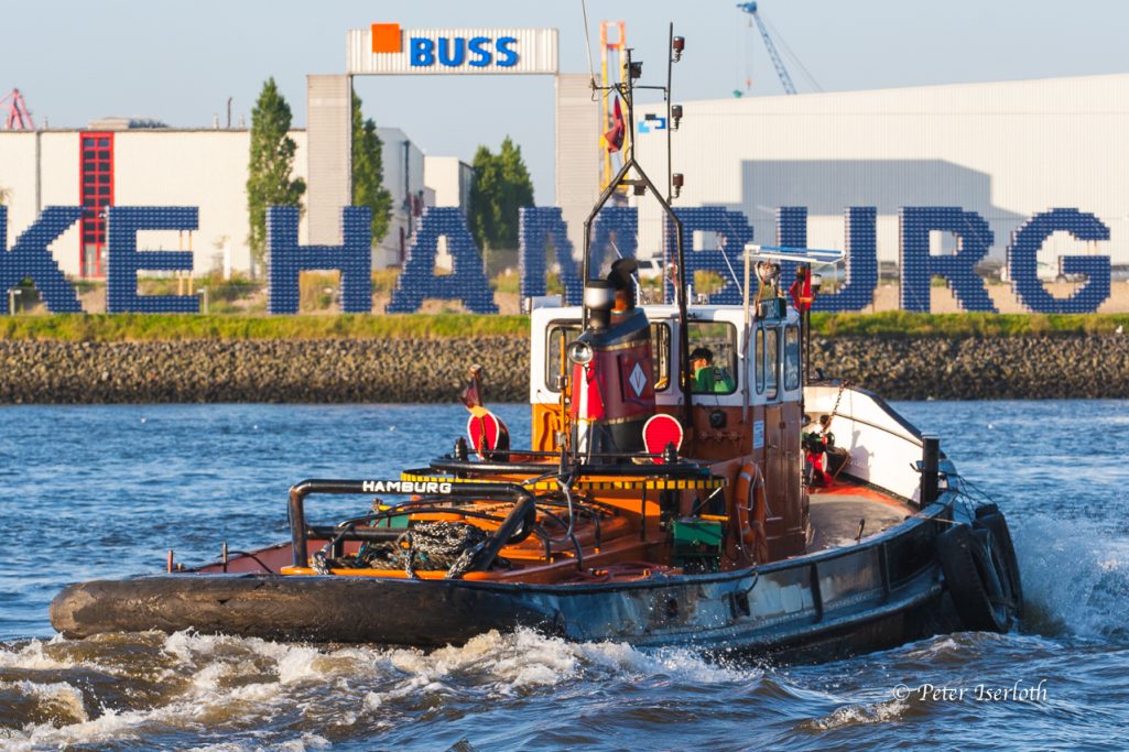 Ein Schlepper fährt auf dem Wasser vor der Hamburger Hafenlandschaft.