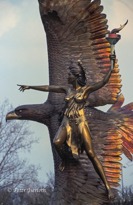 Skulptur einer weiblichen Figur, die eine Fackel hält, mit einem Adler im Hintergrund.