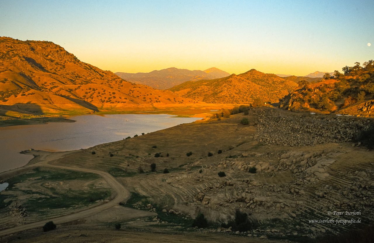 Panorama eines Bergsees bei Sonnenuntergang, umgeben von Bergen und sanften Hügeln.