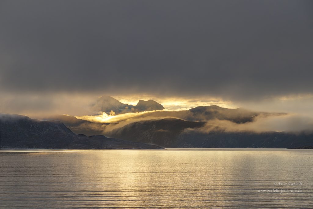 Fotografie eins Sonnenaufgangs , mit dicken Wolken und durch Wolkenlücke, der Blick auf die Berge Grönlands, im Vordergrund ist Wasser.