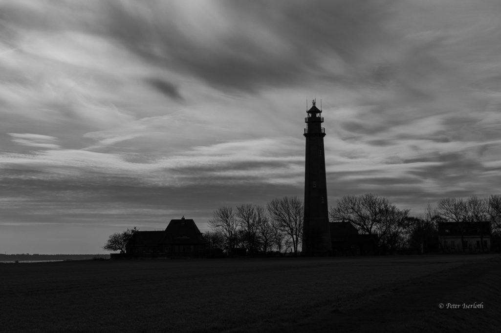 Schwarz-weiß-Anblick eines Leuchtturms vor bewölktem Himmel und silhouettierten Bäumen.