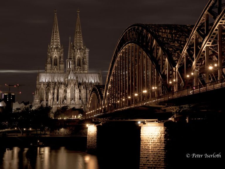 Kölner Dom und Hohenzollernbrücke bei Nacht, beleuchtet am Rheinufer.