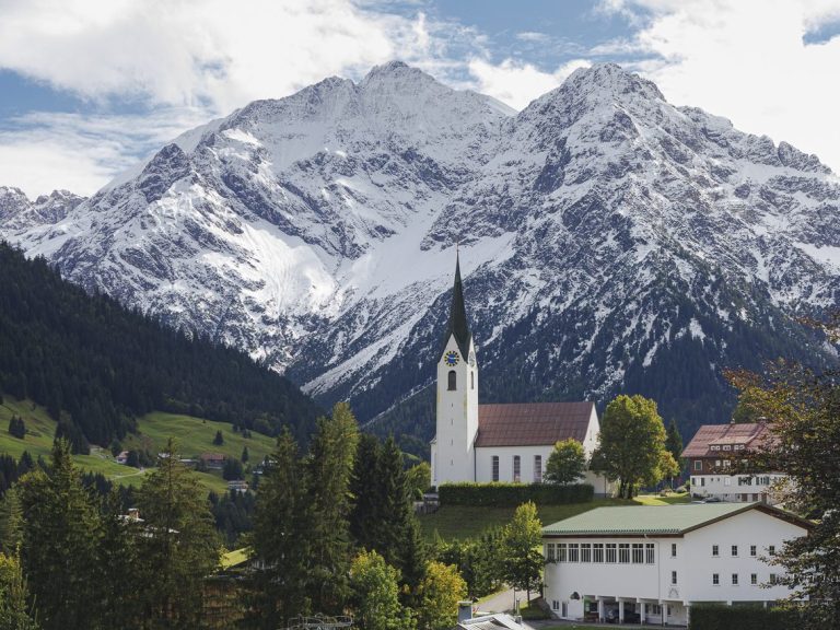 Fotografie der Pfarrkirche hl. Anna Kleinwalsertal, mit Berglandschaft im Hintergrund.